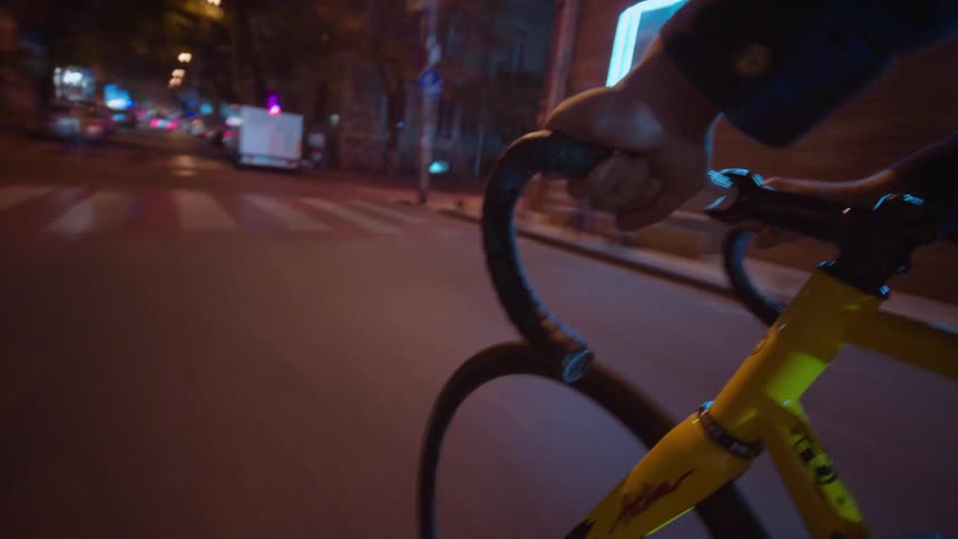 A person is riding a bicycle at night on a city street filmed from a perspective that shows the riders hand gripping the handlebars and the yellow frame below with blurred car lights and illuminated signs in the background
