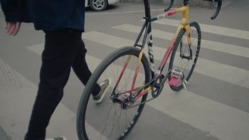 A medium close rear view of a man in a dark jacket and jeans walking a bright yellow and black racing bicycle with red pedals across a zebra crossing in an urban setting