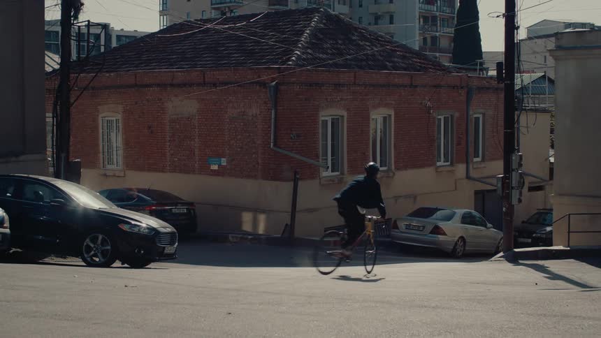 A low angle distant view shows a man on a black racing bicycle quickly riding away on a city street with parked cars passing an old red brick building with a red tile roof in a sunny urban area