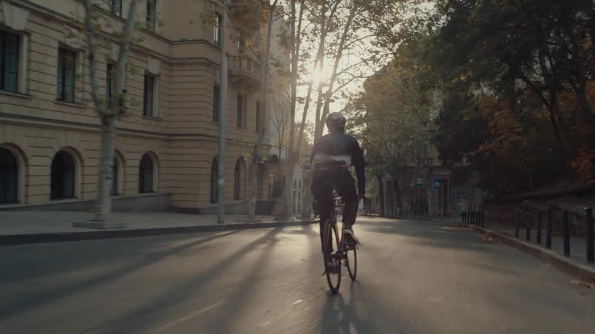 A man rides a bike through city streets at sunrise wearing a helmet as golden light reflects off buildings and pavement