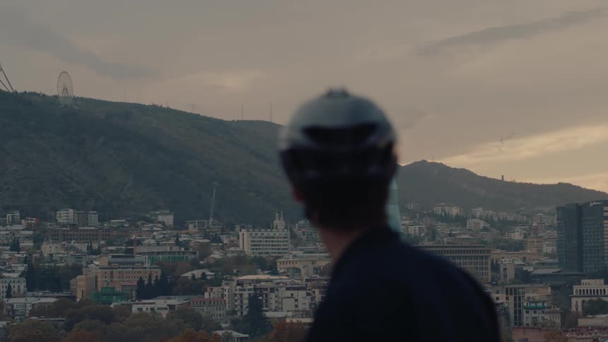 The video shows a mans back and head slightly out of focus looking out over a cityscape at dusk or on an overcast day the scene features a view of a hilly terrain with many buildings and a ferris wheel visible on one of the hills in the backgr