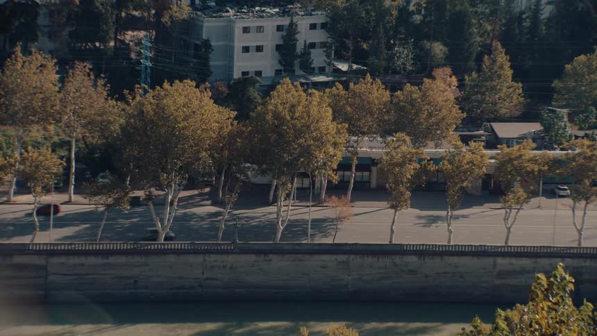 An urban street lined with trees displaying yellowish foliage running parallel to a gray concrete embankment above a river with cars driving and parked on the street and multistory buildings visible on the hillside in the background
