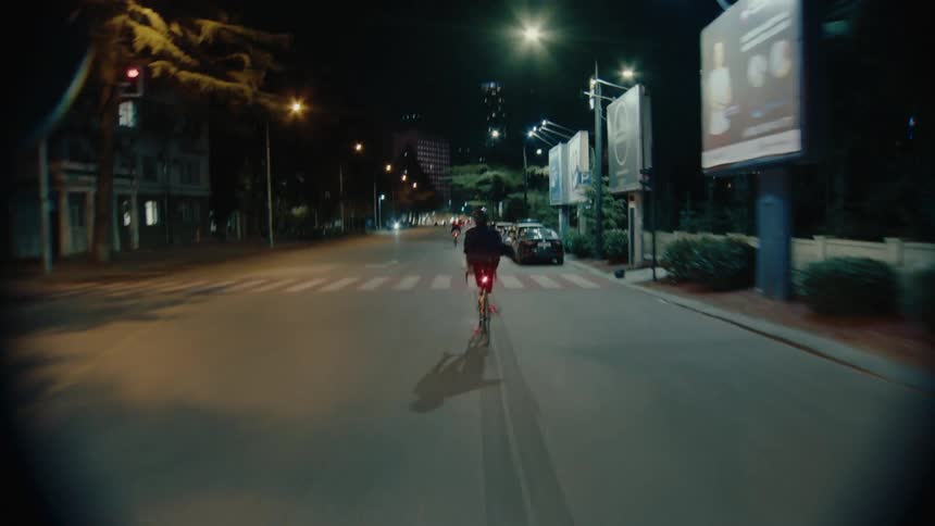 A moving follow view of several cyclists riding racing bicycles on a wide city street at night with bright yellow markings and red tail lights visible in the dark urban setting