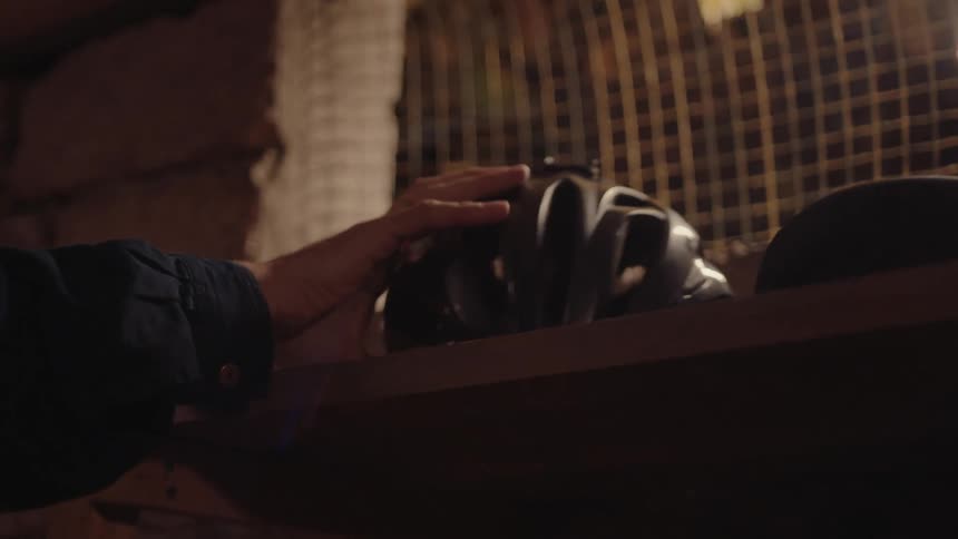 A mans hand reaches up and quickly grabs a black bicycle helmet from a dark wooden shelf against a stone wall and wire mesh background in a dimly lit basement or storage area