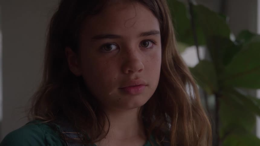 Girl standing indoors near the window turns her head to stare sadly