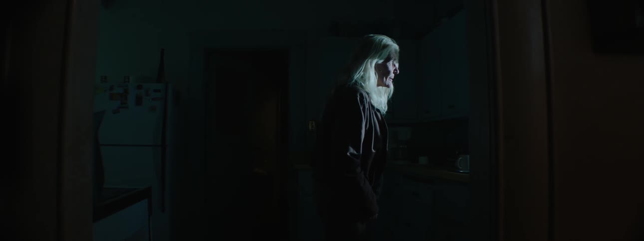 A woman walks into a dark kitchen and looks out the window