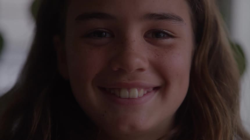 The video is a closeup shot of a young girls face smiling naturally and looking off to the side she has freckles and long brown hair and the lighting is soft and natural