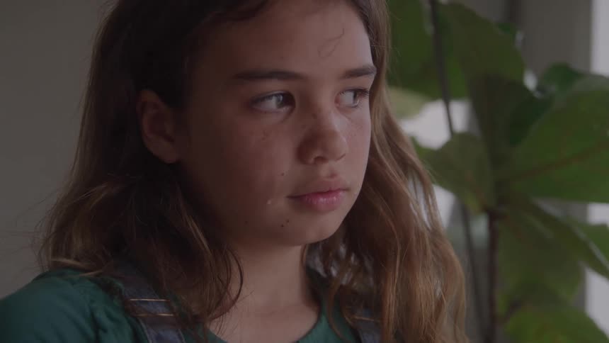 Girl with a serious expression looks to the side near a window with plants in the background