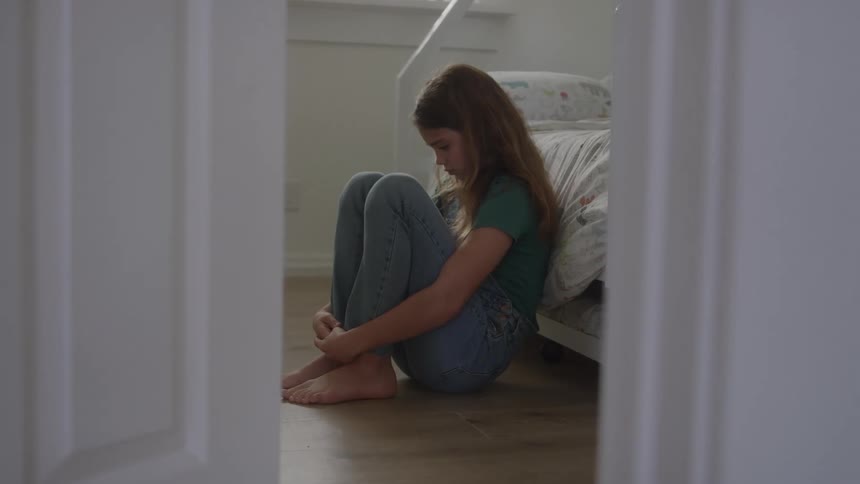 A side view framed by an open doorway shows a young girl with long brown hair sitting alone on the wooden floor with her knees pulled to her chest looking sad and isolated in a bright bedroom