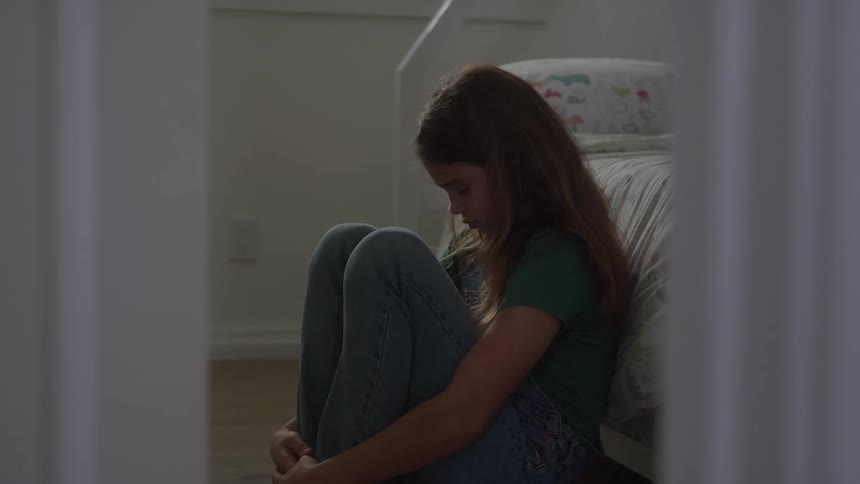 A girl sadly sits against her bed