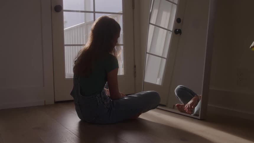 Teenager girl wearing a denim jumpsuit sitting on a wooden floor looking at the mirror and down in a depressed manner indoors during day time