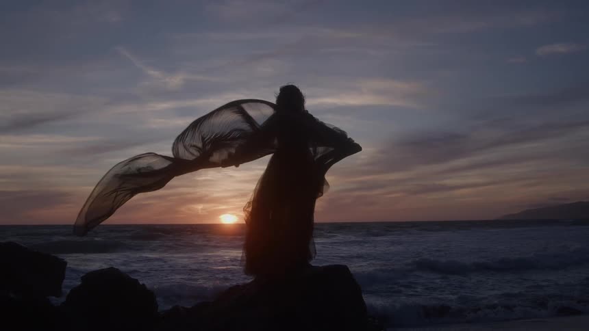 Woman with flowing fabric standing before the sunset by the sea
