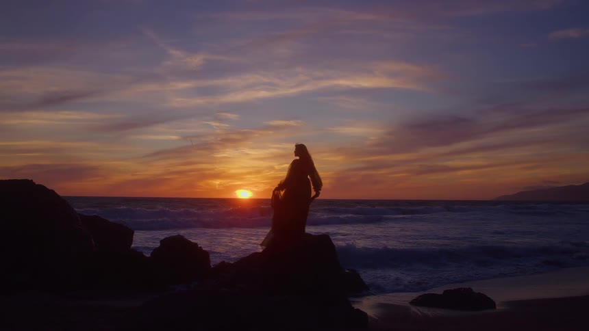 A woman silhouette on a large rock at the edge of the ocean holding sheer material that is briefly illuminated by the low sun positioned directly behind the person as waves break against the shore under an orange and purple sky