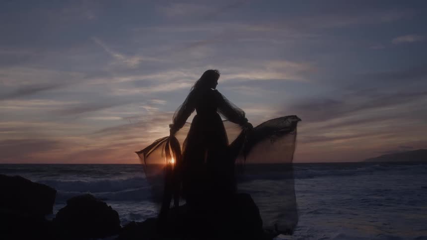The video shows the silhouette of a person in a dress and veil standing on dark rocks by the ocean at sunset the sky is a dramatic mix of orange and purple highlighting the figure and their flowing attire against the waves