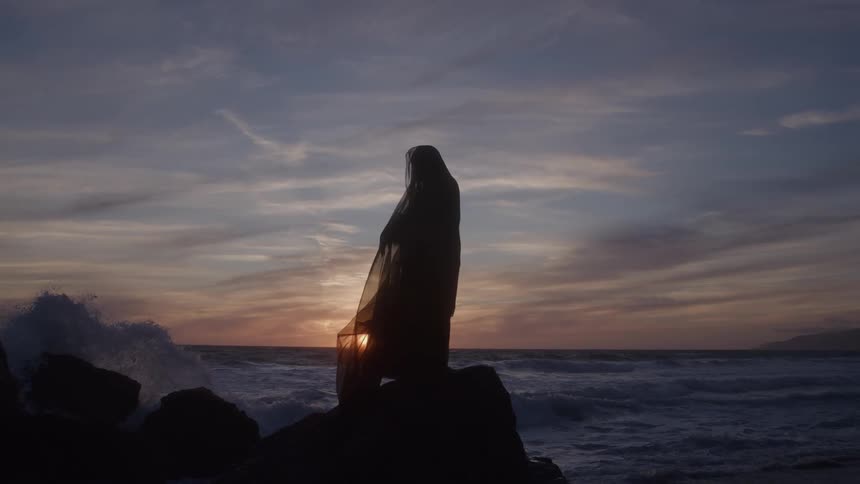 The woman in a black dress stands on the rocks of the shore
