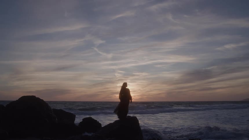 The video shows the silhouette of a person in a flowing dress or robe standing on dark rocks by the ocean at sunset the sky is filled with a striking blend of orange pink and purple clouds creating a dramatic backdrop against the figure an