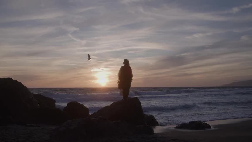 The video shows a person in a flowing dress or robe silhouetted against a sunset on a rocky beach the sky features a mix of warm sunset colors and high clouds providing a dramatic backdrop for the figure standing on the dark rocks