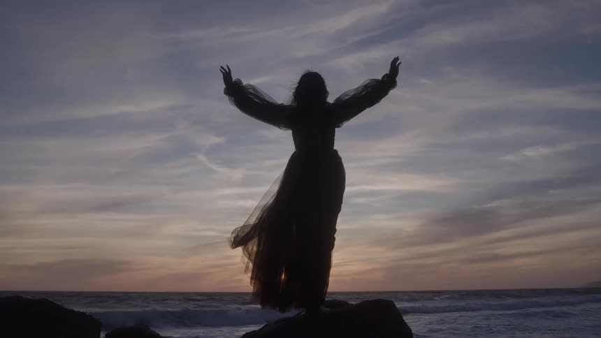 Silhouette of a woman with open arms facing the sea at sunset