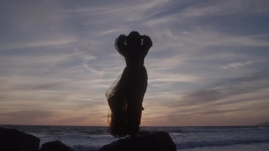 Silhouette of a woman raises her arms facing the sea during sunset