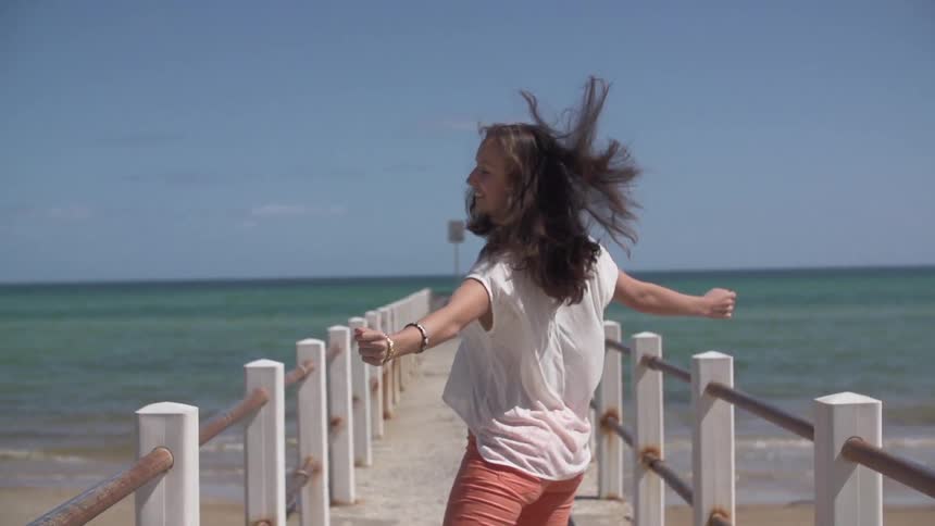 A young woman with long hair smiling and twirling around with her arms out as she walks down an ocean pier