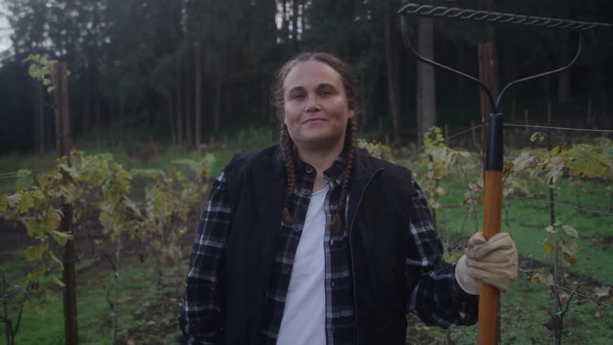 Woman with braids holds a rake in a vineyard at dusk