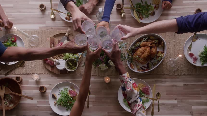 An overhead view shows six people sitting around a long wooden table set for dinner all raising their glasses in a toast over plates of salad and a roasted chicken with a charcuterie board and a floral centerpiece in the middle
