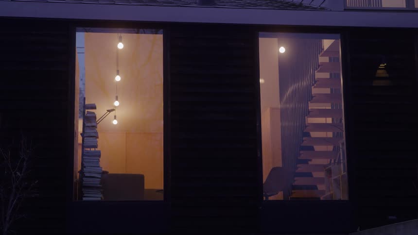 The video is a stationary long shot of a modern twostory house at night with large rectangular windows the windows reveal a warm illuminated interior featuring a stack of books and dangling light bulbs in the left frame and what appears to