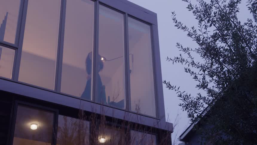 The video is a lowangle long shot of a modern square house with large windows against a dusk or overcast sky a person is vaguely visible as a silhouette through the upper window suggesting they are standing indoors and looking out