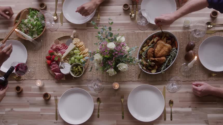 The video is an overhead shot of a table set for a dinner party with multiple peoples hands visible the table is filled with food including a roast chicken a cheesecharcuterie board and a bowl of salad alongside plates cutlery and wine gl