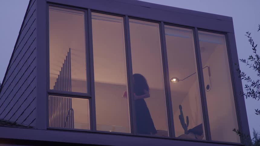 The video is a stationary medium shot of a modern square building with large tall windows inside the window a womans silhouette is visible against the light and a stack of books and a string of lights are also noticeable