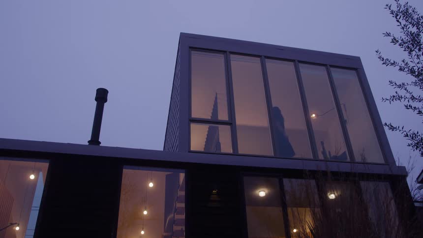 A medium low angle exterior shot shows the second floor of a modern dark house with floor to ceiling windows reflecting the twilight sky and a woman in a dress walking inside with string lights glowing warmly indoors