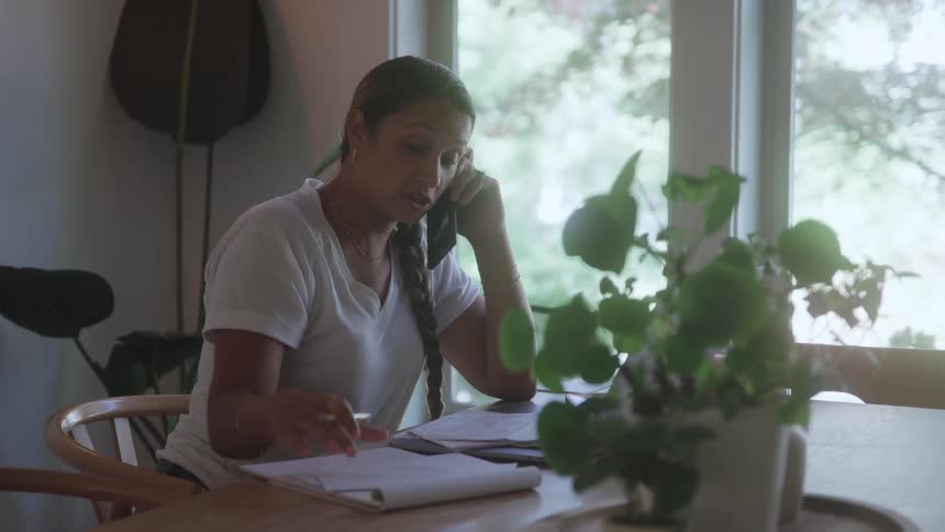 A young woman sits at a dining table at midday sorting through mail documents while talking on the phone focused and multitasking as sunlight filters into the room