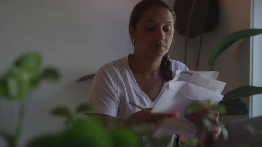 A young woman sits indoors at midday surrounded by potted plants as she sorts through a stack of mailing documents her tired expression reflecting the weight of the task