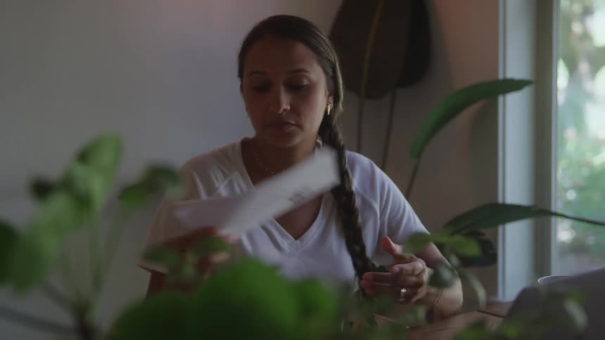 Woman sits with her hands on her head near a sunlit window with plants