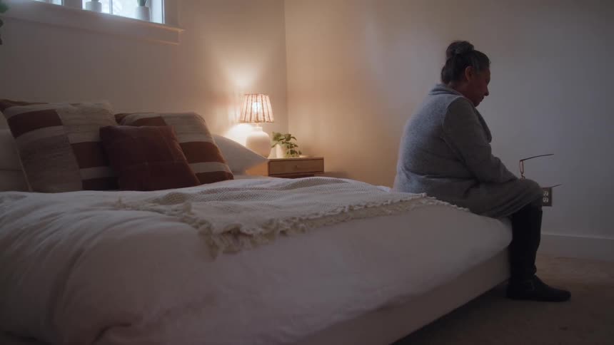 Elderly woman sitting on the corner of a bed in a living room looking down and depressed indoors during day time