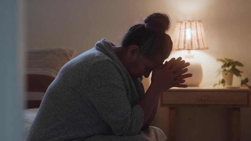 The video is a stationary medium shot showing a woman seated in a room illuminated by a warm lamp light on a bedside table she is seen in profile with her head bowed and hands clasped suggesting a moment of quiet reflection or prayer