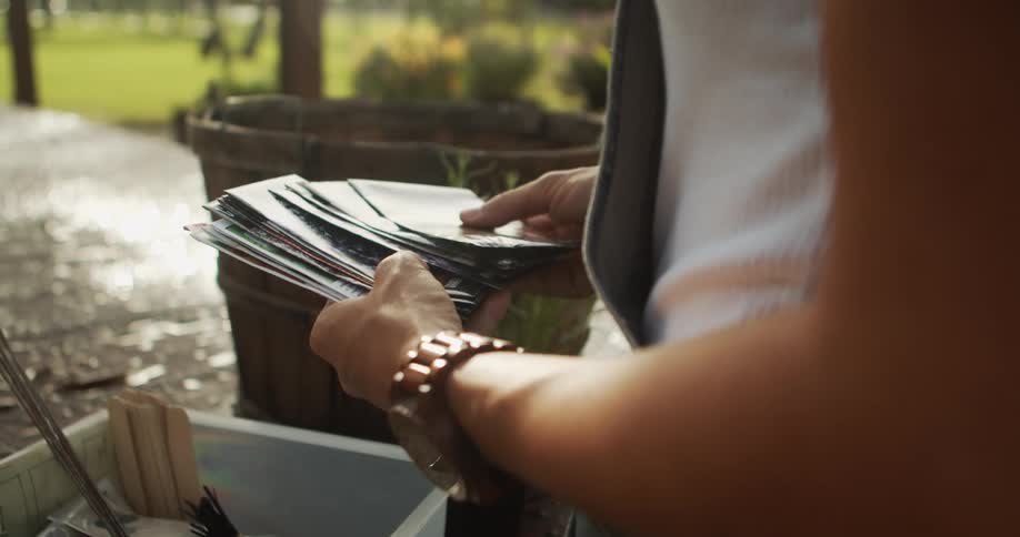 A close view shows a person with a braided bracelet wearing a white shirt sorting through a stack of gourd seed packets with a wooden barrel in the blurred background