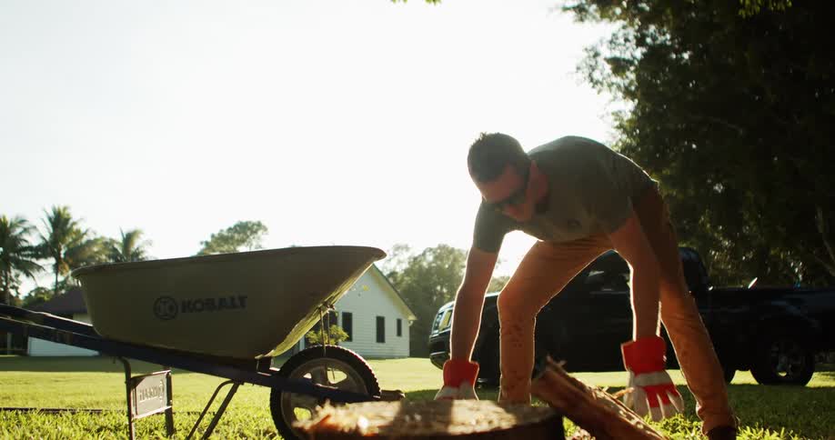 A man wearing sunglasses a gray shirt and orange gloves loads split firewood into a yellow wheelbarrow on a green lawn with a white house and bright sun flare in the background