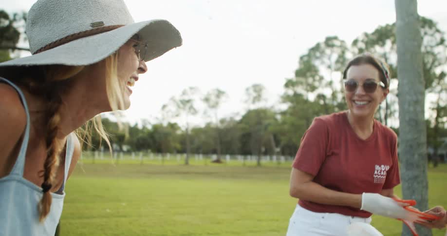 The video shows a midshot of two women in an outdoor grassy area both smiling and looking at the person offcamera one woman wears a widebrimmed sunhat and denim overalls while the other wears a red tshirt and sunglasses