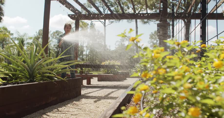 The video is an outdoor shot featuring a woman spraying a garden with a hose nozzle the scene is bright and sunny showing raised garden beds and potted plants under a wooden pergola