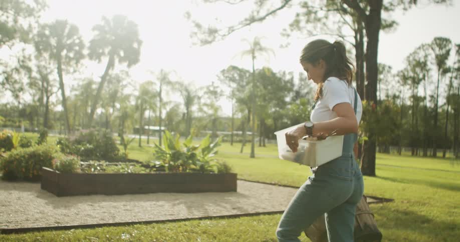 The video shows a woman walking away from the camera in a grassy sunny outdoor area carrying a storage box and a backpack she is wearing a white tshirt and denim overalls and the background suggests she is in a large yard or farm setting