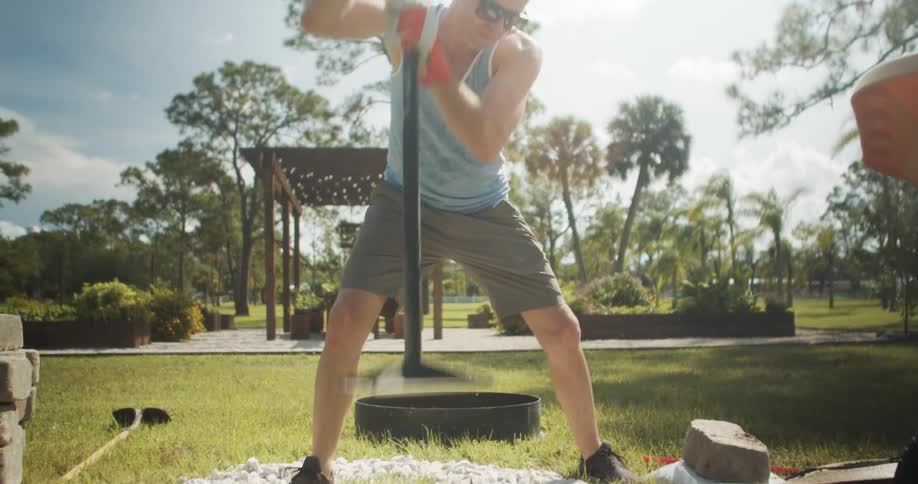 The video shows a man in a sleeveless shirt using a posthole digger in a sunny grassy backyard in the background there is a wooden pergola structure and trees suggesting a home garden or outdoor project