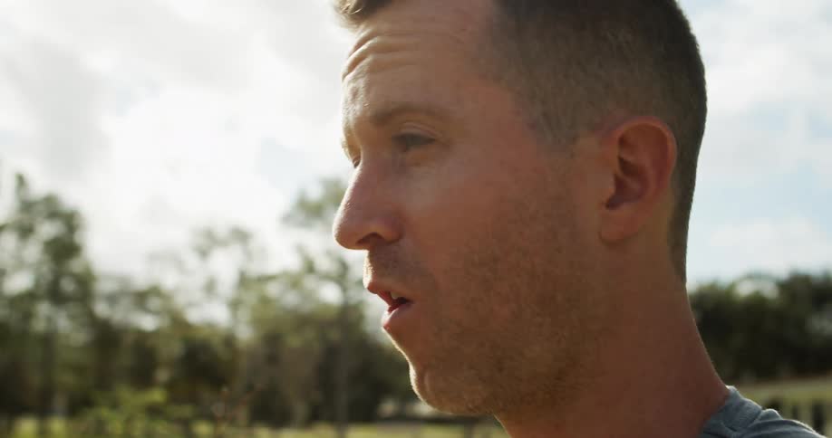 A side profile close view shows a man with short brown hair closing his eyes and inhaling deeply while bending over a plant with green leaves outdoors on a sunny day