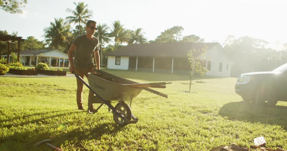 Person pushes wheelbarrow across lawn in residential area with palm trees and houses in the background