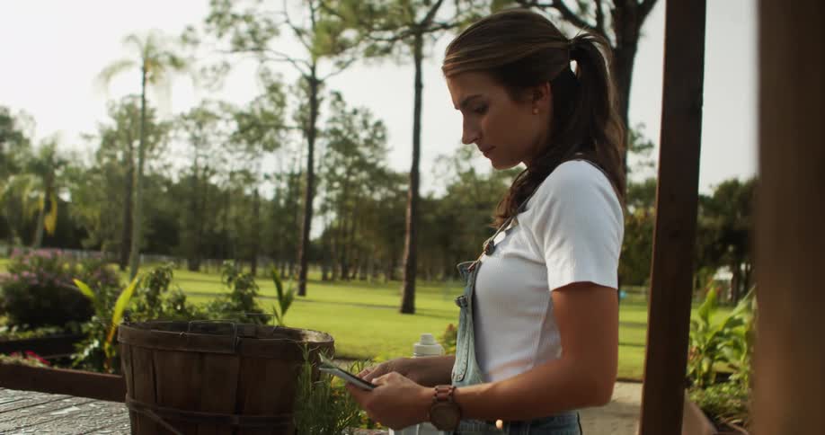 The video is a midshot of a young woman with her hair in a ponytail standing outdoors in a garden area during the day she is wearing denim overalls and a white shirt and is looking down handling small seed packets