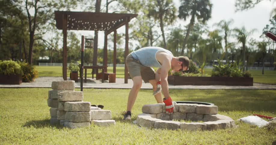 The video is an outdoor shot featuring a man constructing a circular fire pit in a grassy backyard he is wearing gloves and placing large stacked paving stones while working near a wooden pergola structure