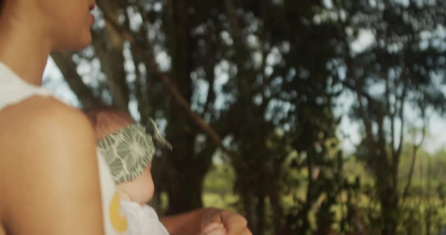 Young mom holding her baby daughter and playing with her hand while smiling next to trees and vegetation outdoors during day time