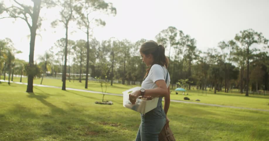 Woman walks across lawn carrying white box and brown basket in residential area with houses in the background
