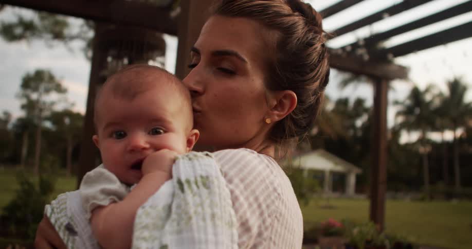 The video is a mediumcloseup of a smiling woman holding a baby outdoors both the woman and the baby are looking directly at the camera and the background suggests they are in a grassy yard near a wooden pergola structure