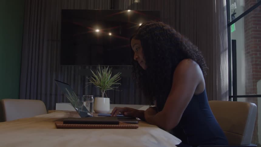 The video is a midshot of a woman with long curly hair sitting at a table in a modern office or cafe setting she is attentively working on a laptop and writing on a tablet or notebook